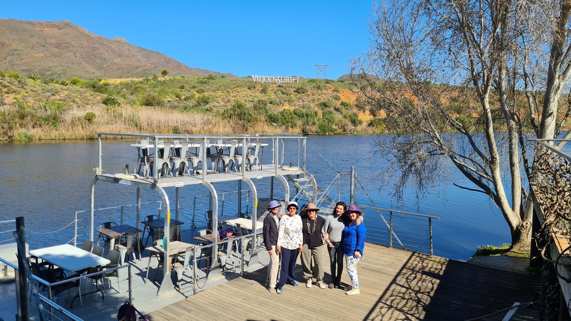 Group of people on a dock by a lake with a wakeboarding structure, mountains in the background.