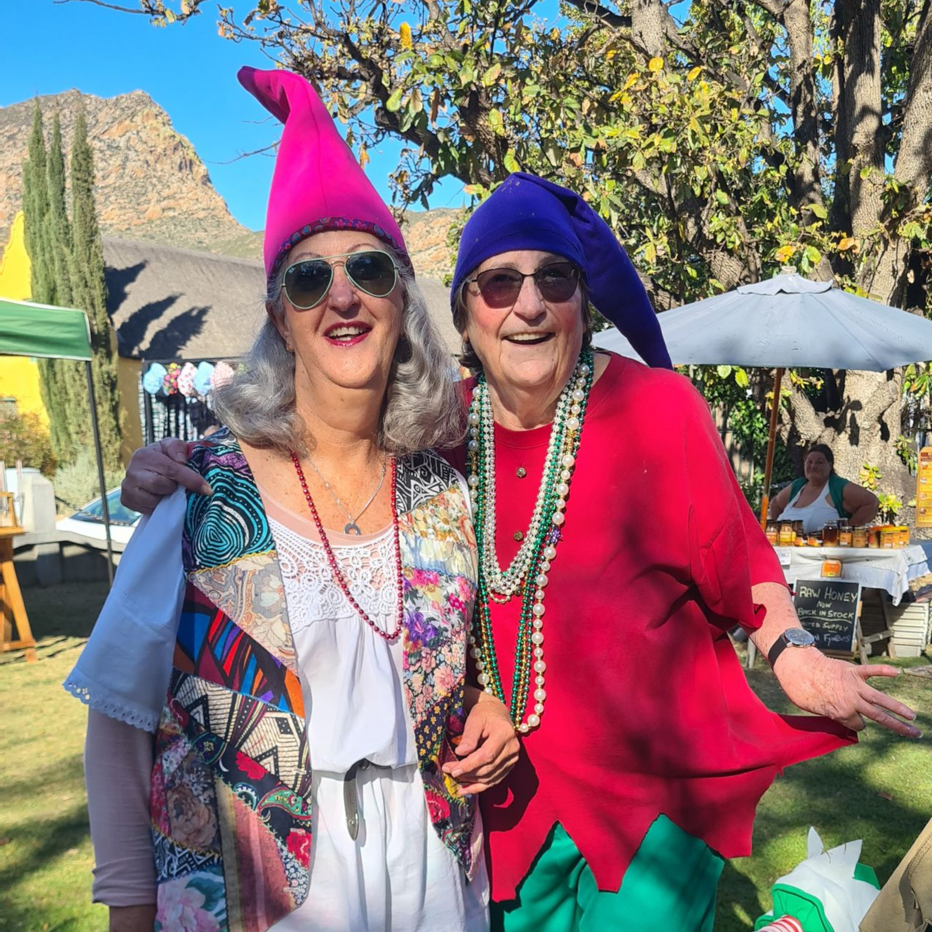 Two women smiling, wearing gnome hats, outdoors. One in white, patterned vest, the other in red.