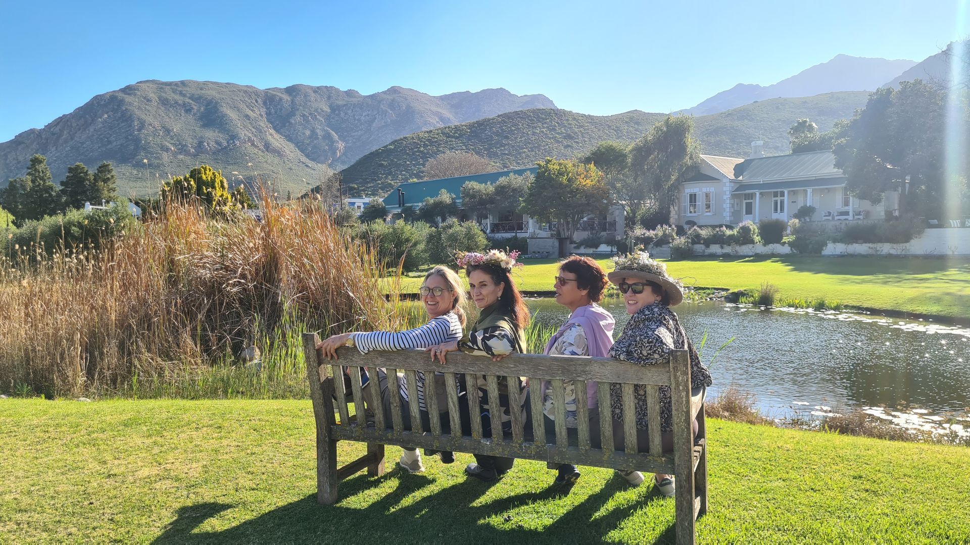 Four people seated on a bench, overlooking a pond, green lawn, and mountains on a sunny day.