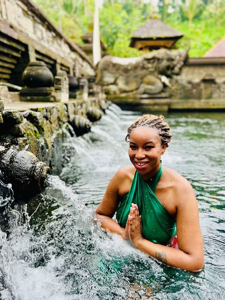 Woman in green wraps, in water, hands together, smiling at Tirta Empul Temple, Bali.