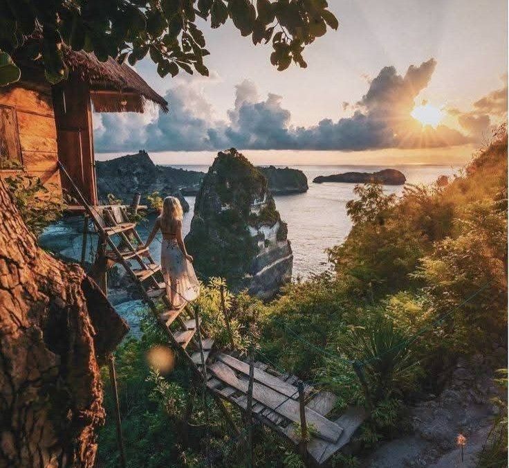 Woman on a wooden walkway toward a seaside cliff with a hut at sunset.