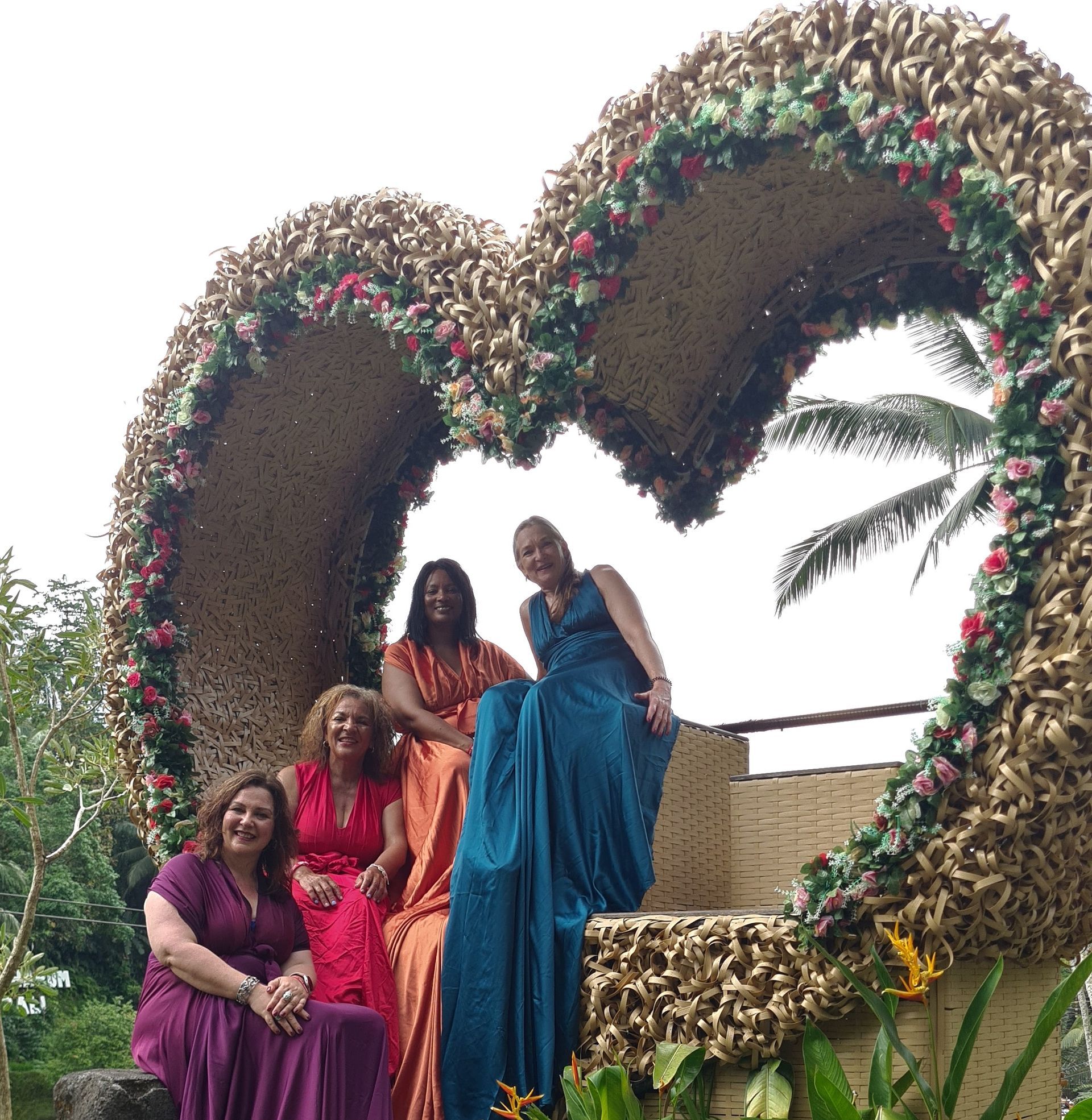 Four women in colorful gowns pose in a large heart-shaped floral arch outdoors.