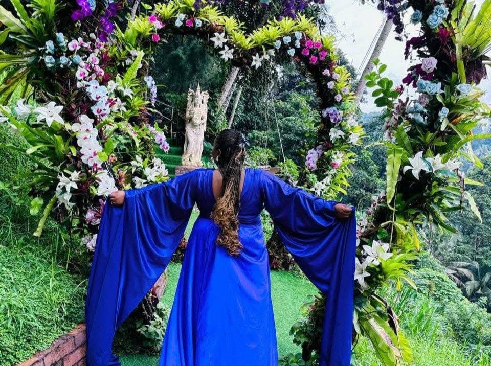 Woman in blue dress stands under floral heart archway, arms outstretched, outdoors.