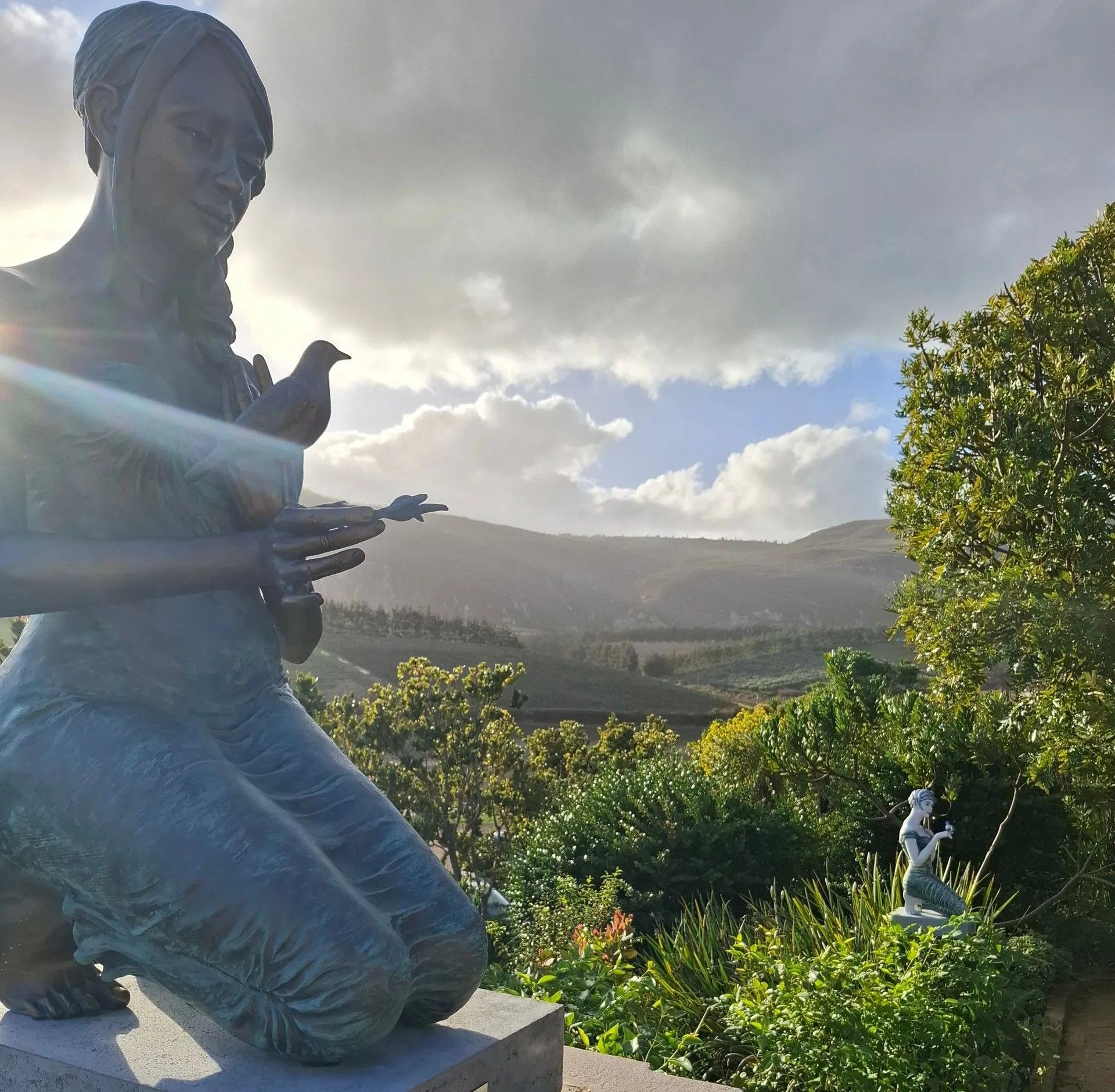Bronze statue of a woman kneeling with doves, mountains and sunny sky in the background.