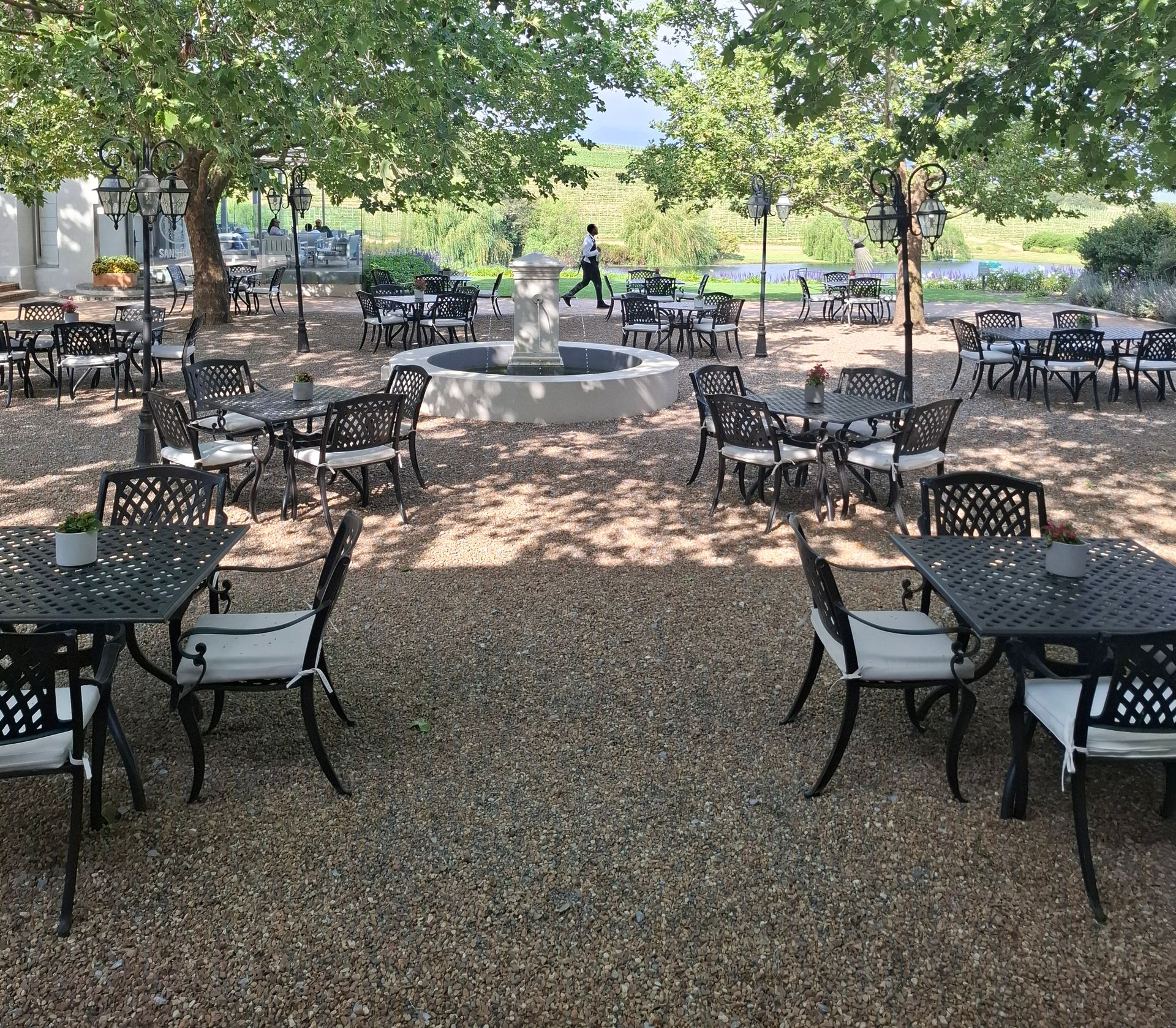 Outdoor restaurant with tables and chairs under trees, small fountain, person walking.