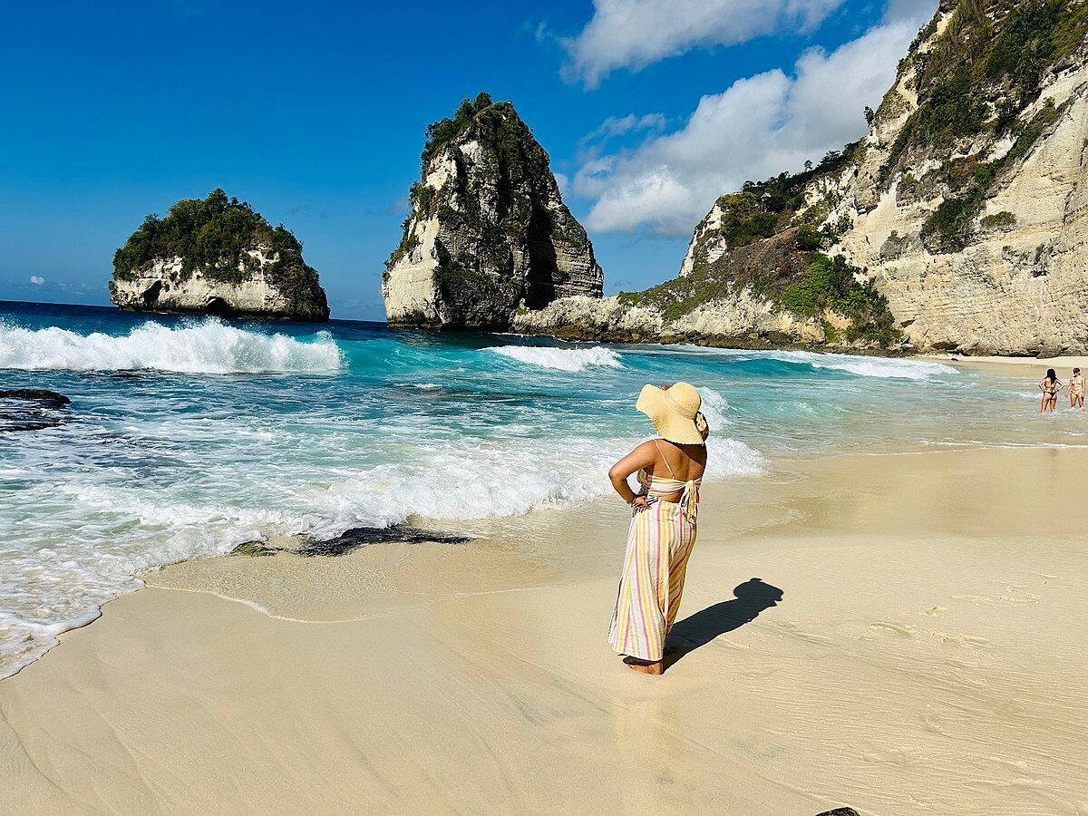 Woman in hat on beach, facing turquoise water and large rock formations under blue sky.