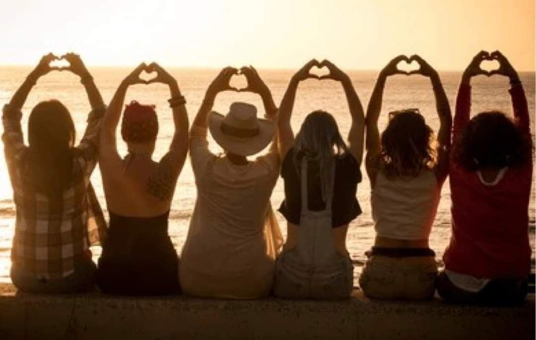 Six women seated, facing sunset, making heart shapes with their hands.