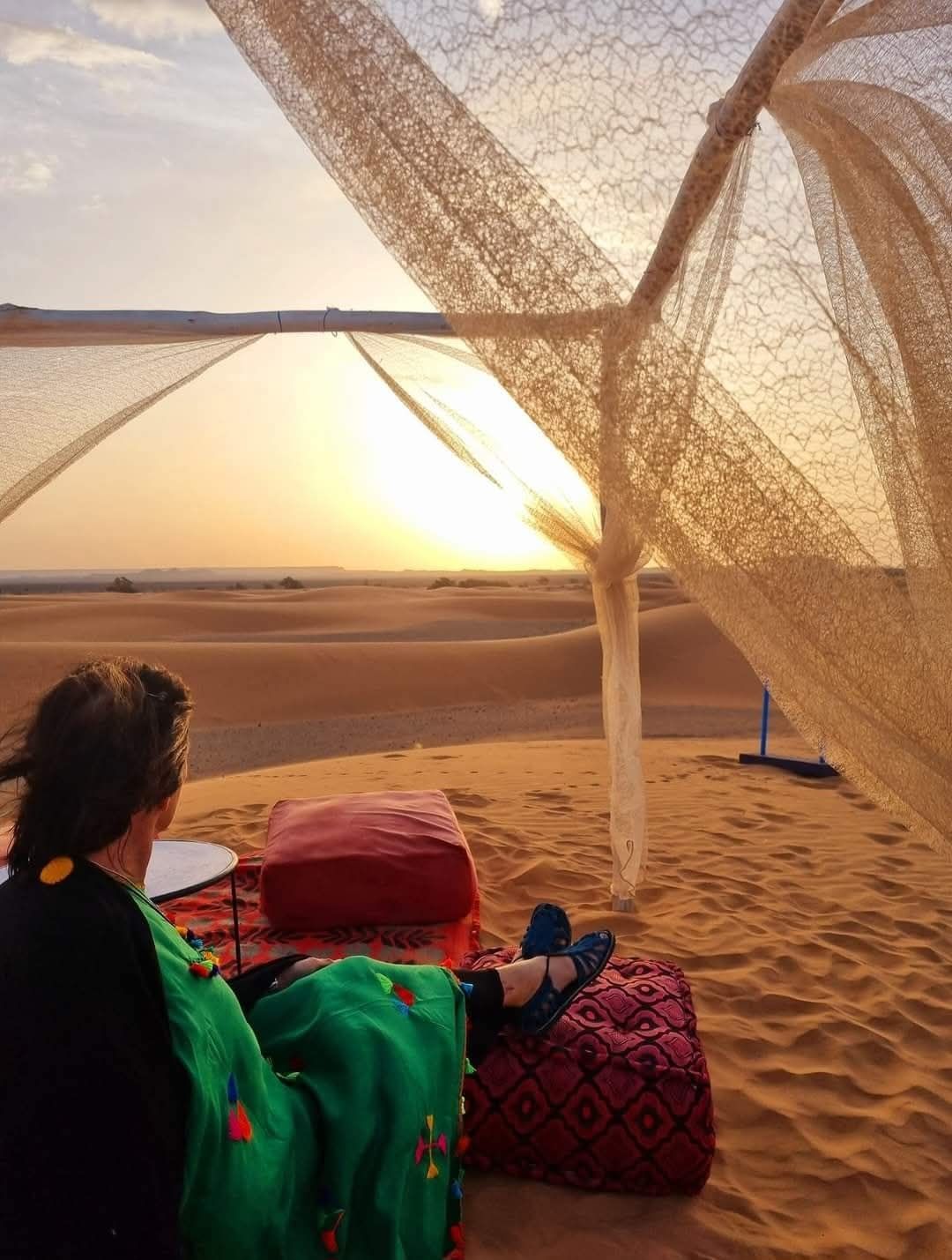 Person lounging on cushions in a desert tent at sunset, with sheer fabric draped around the view.