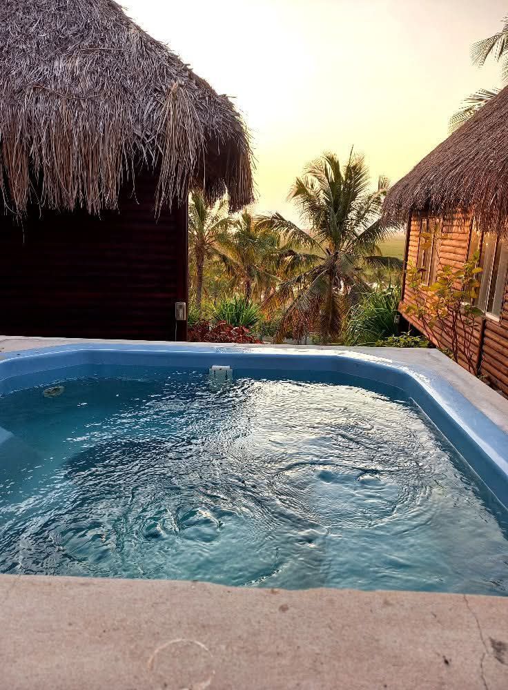 Swimming pool with rippling water, thatched roof huts, palm trees, and sunset.