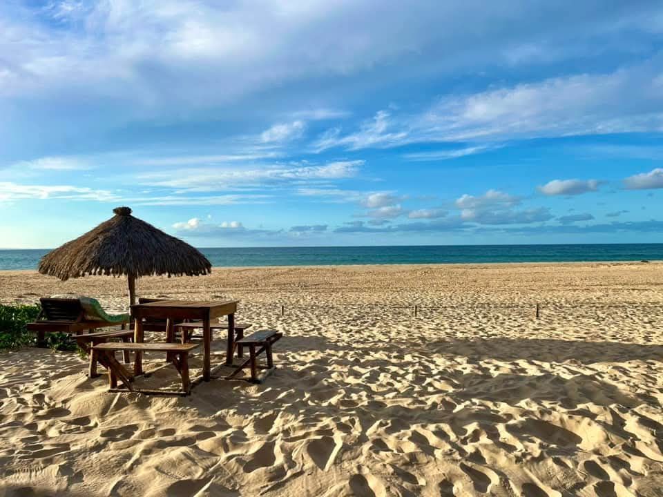 Beach scene: table and chairs under a thatched umbrella, ocean and sky in background.