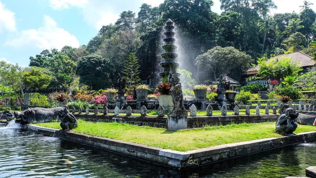 Water garden with fountain, stone structures, and lush greenery in a park-like setting.