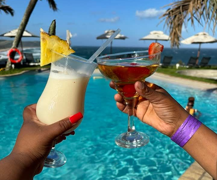 Two hands clink cocktail glasses by a pool with the ocean view. One drink is a piña colada, the other a margarita.