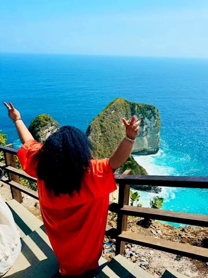 Person with arms raised, overlooking turquoise water and rocky cliffs from a wooden deck.