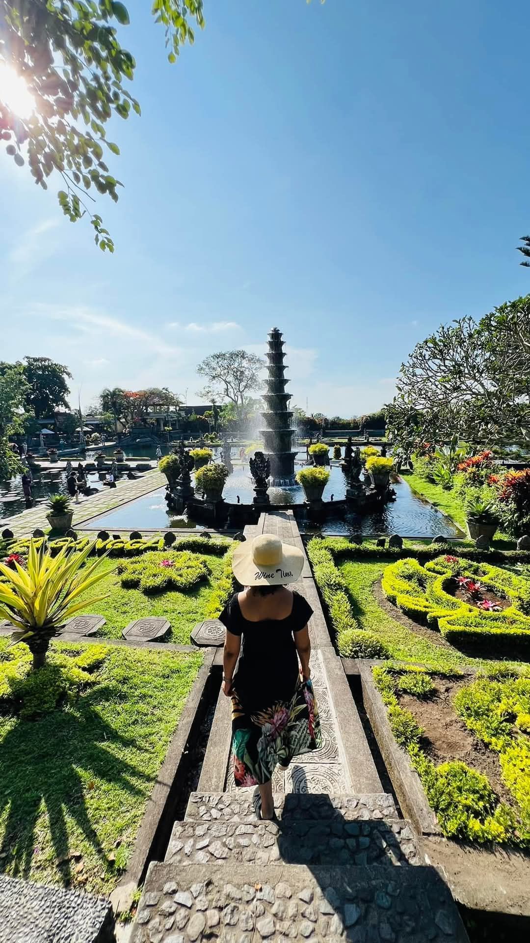 Woman in hat walks down steps toward a fountain in a lush garden, Bali.