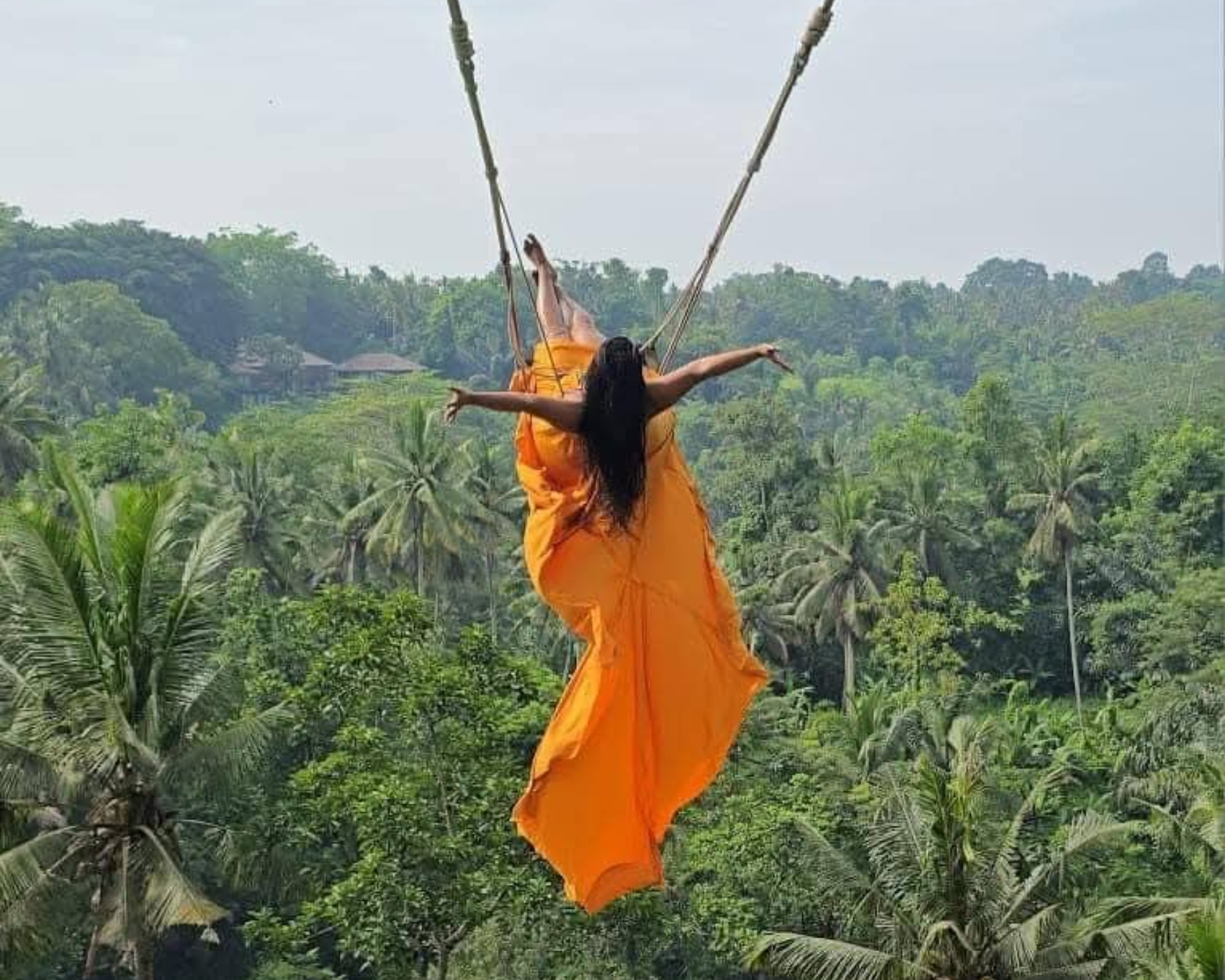 Amarantine Travels' guest in orange dress on swing, arms outstretched, over lush green jungle in Bali.