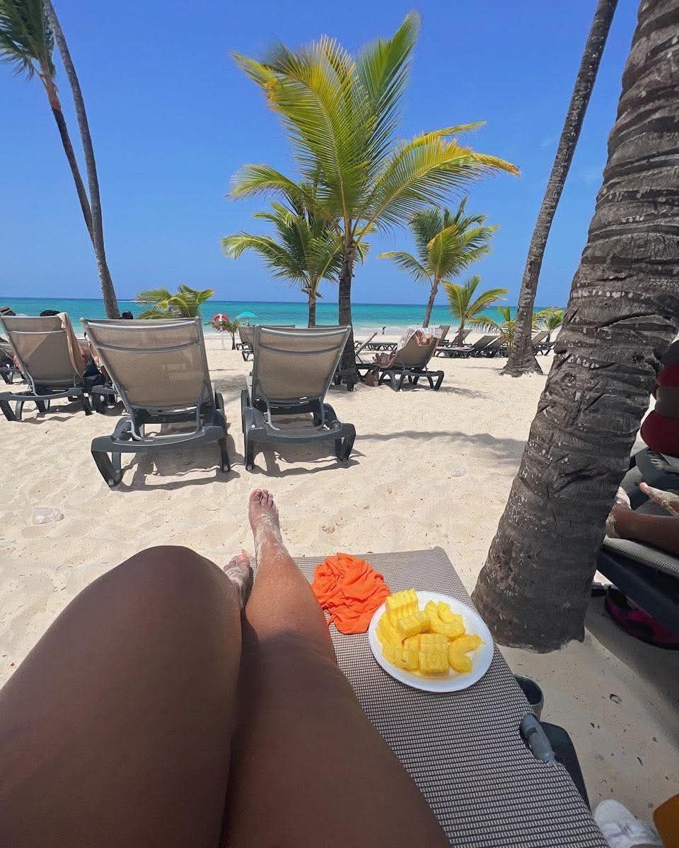 Person relaxing on a beach chair, feet up, eating mango. Turquoise water, palm trees, and a sunny sky.