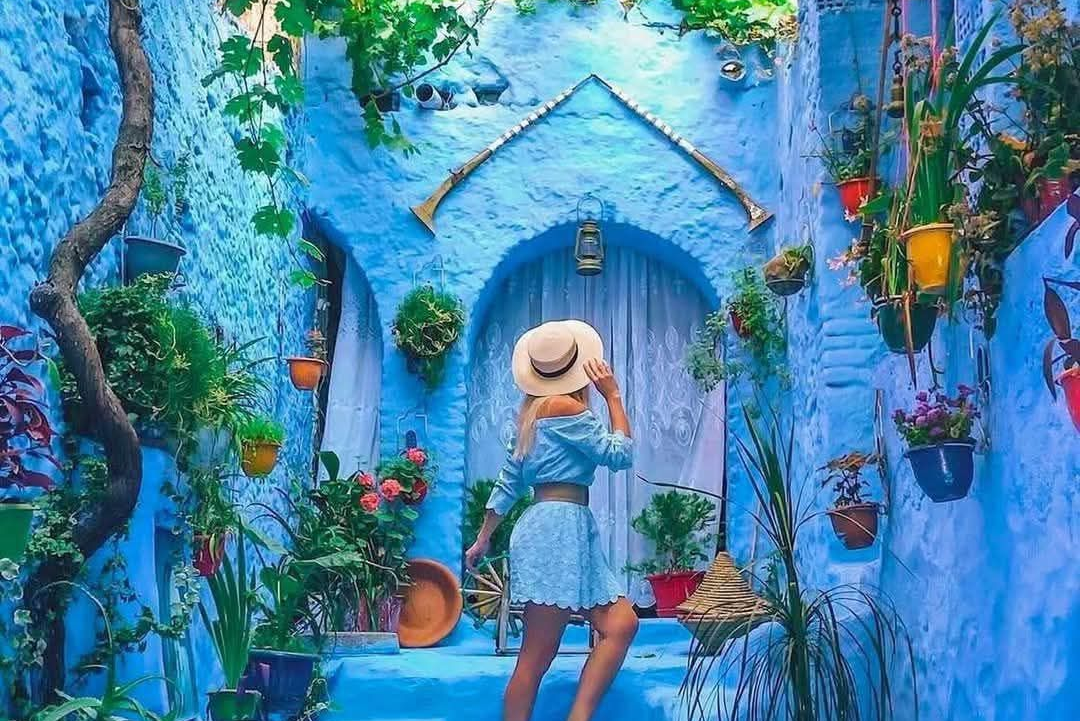 Woman in blue outfit and hat stands in a blue alley with plants and doorway.