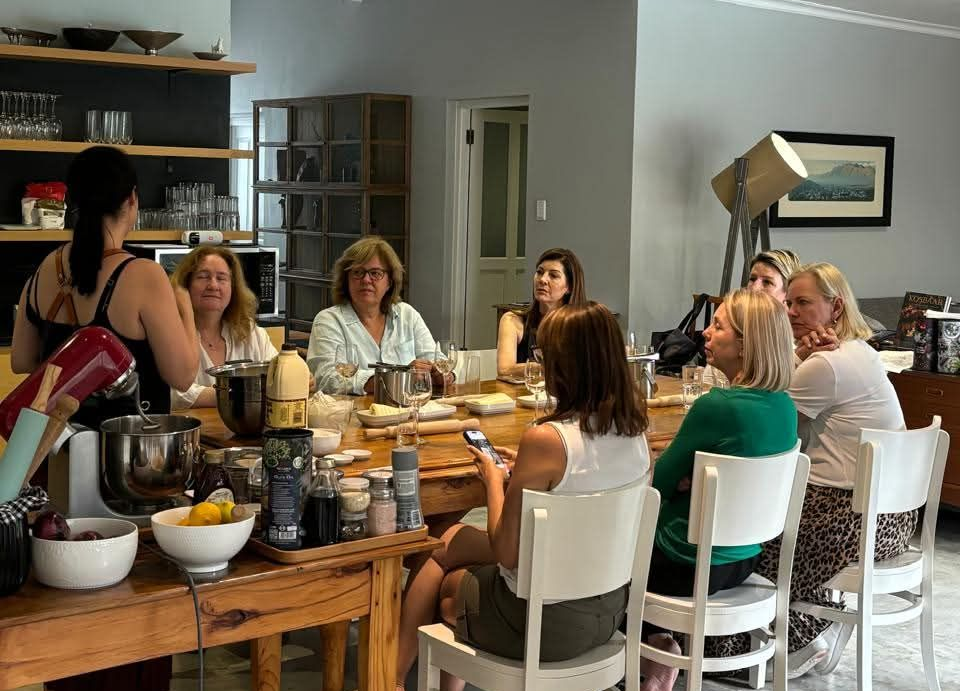 Group of women seated at a table in a kitchen setting; one woman is instructing the group.