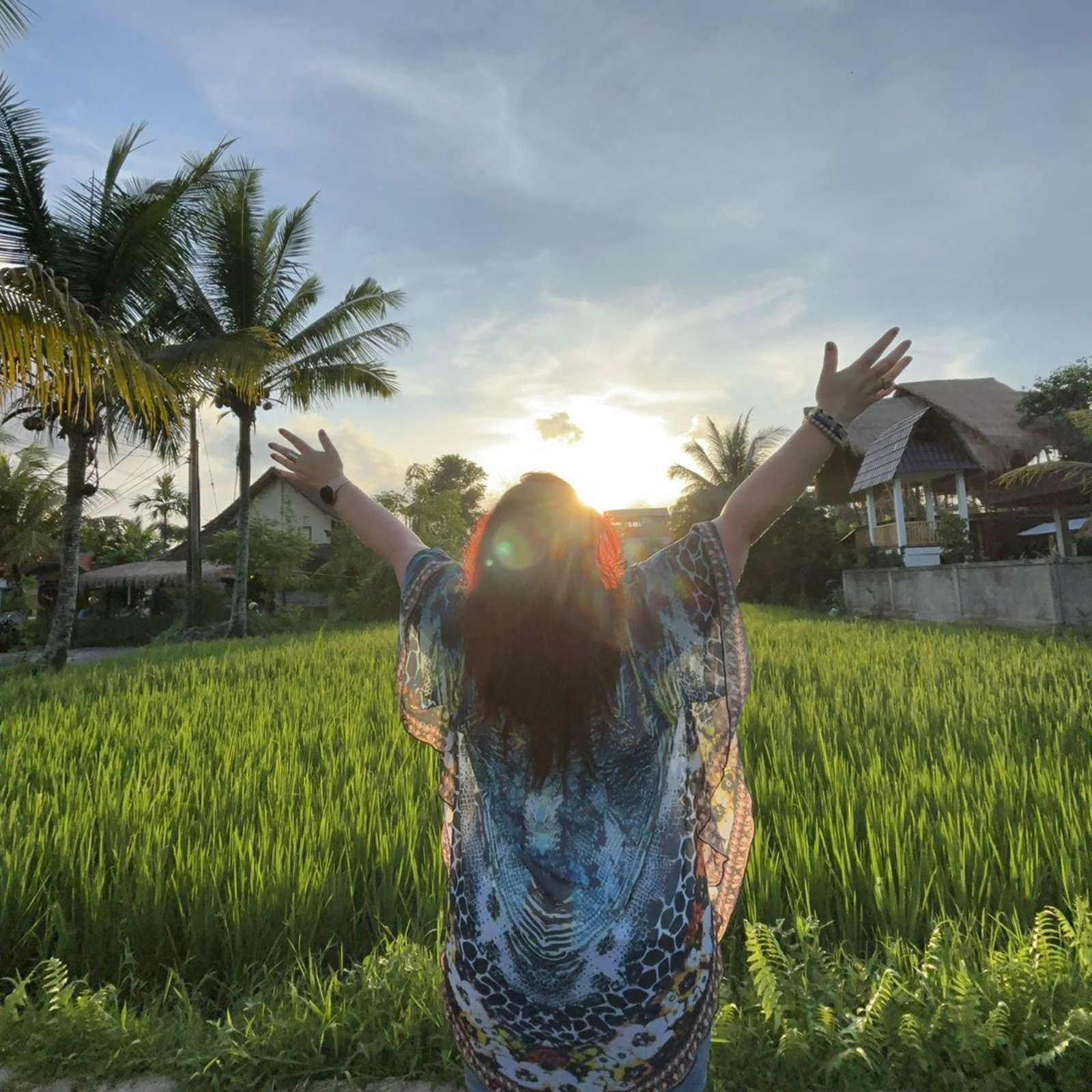Woman with arms raised, facing sunset over rice field, Bali.