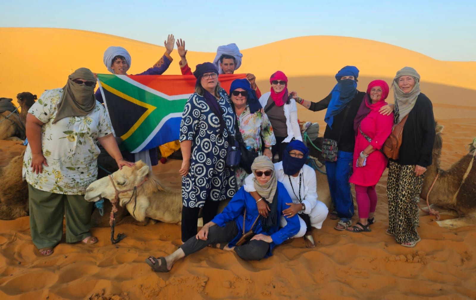 Group of people pose with a South African flag and camels in a desert setting.