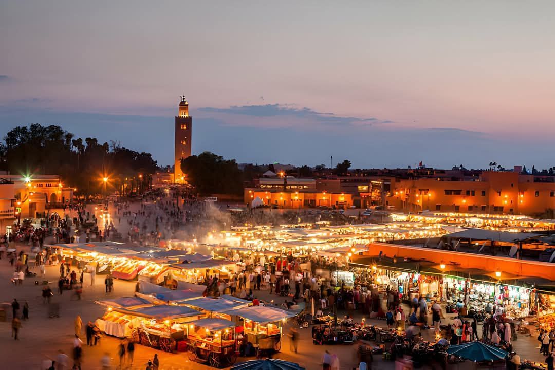 Bustling sunset market square with warm lights, crowds, and a tall clock tower under a pink sky