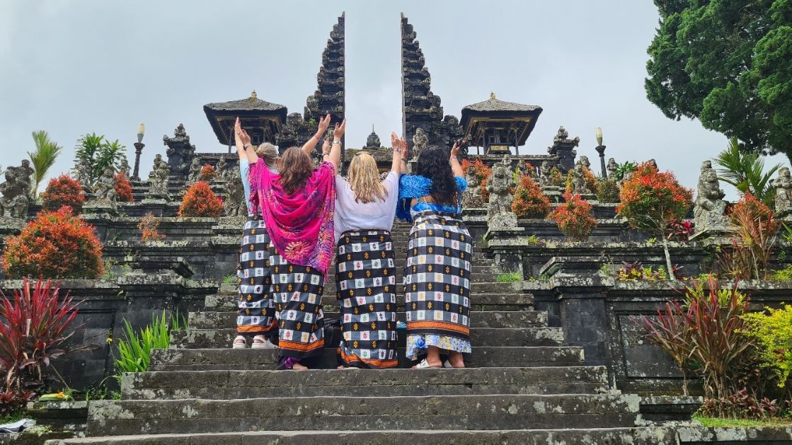 Four people stand on stone stairs facing a traditional Balinese temple gate, hands raised toward the sky.