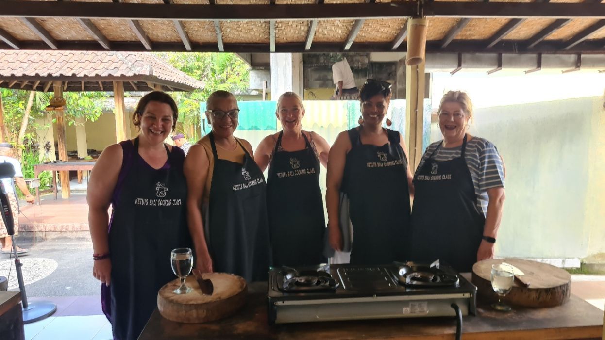 A group of seven people stands smiling on a wooden deck by a pool, with mountains and trees in the background.
