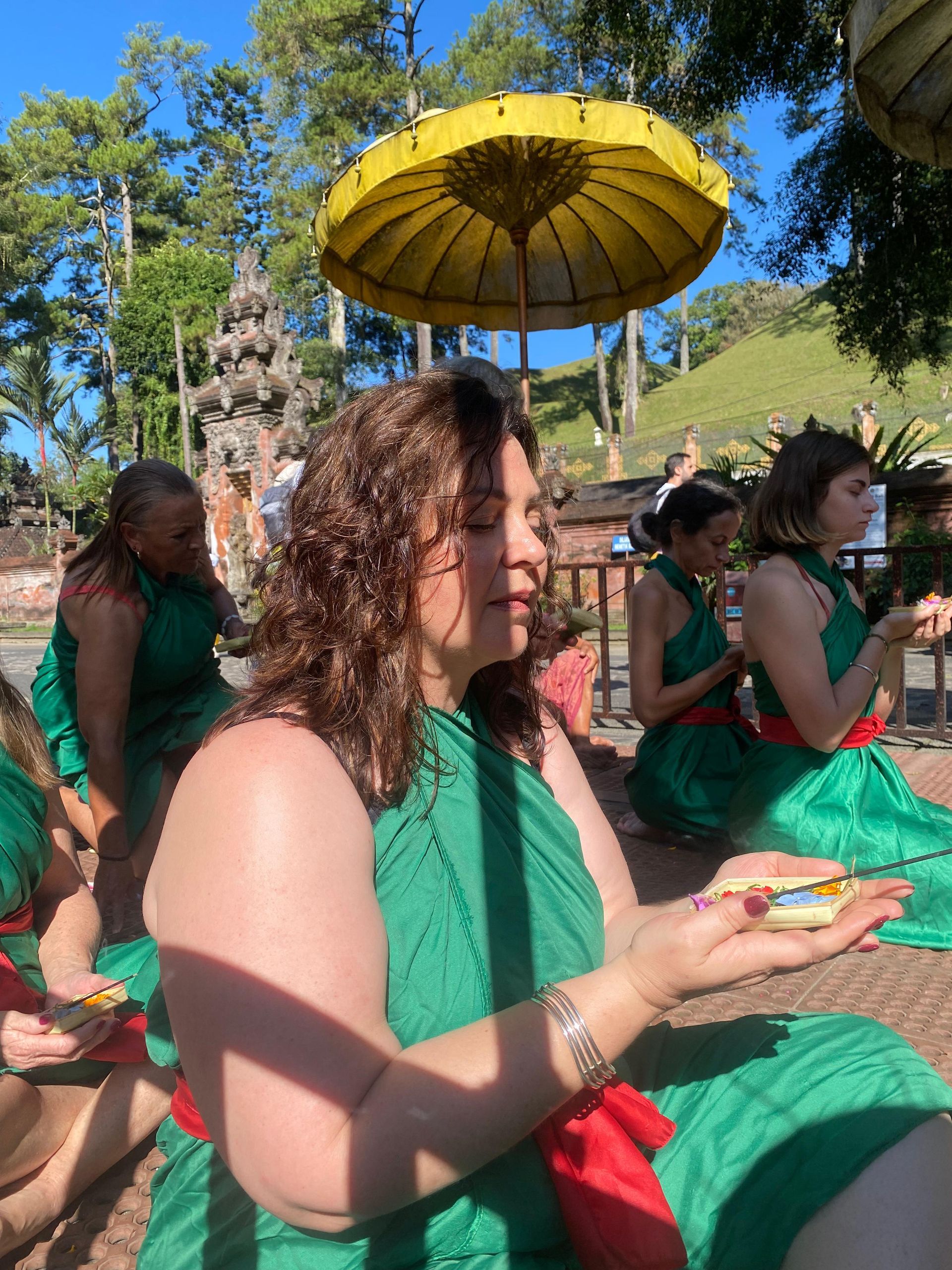 People in green ritual attire with red sashes pray outdoors under a large yellow ceremonial umbrella.