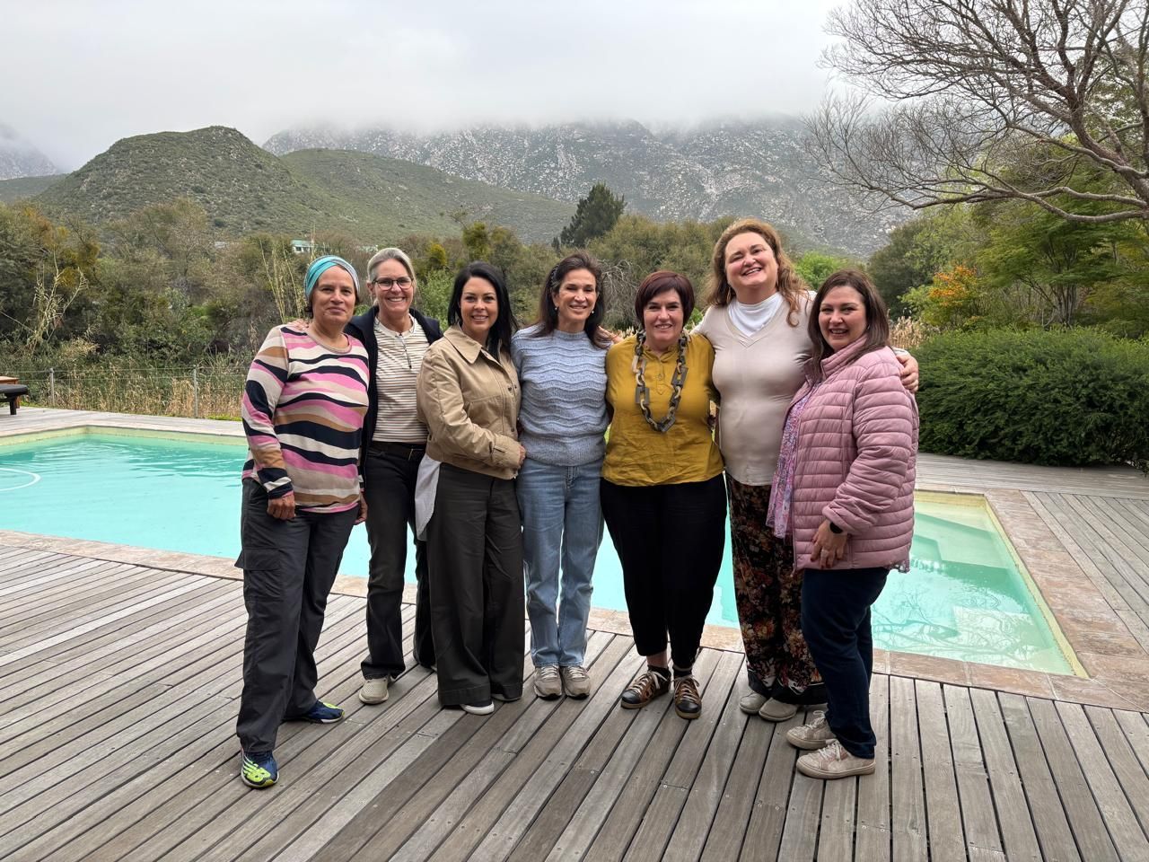 A group of seven people stands smiling on a wooden deck by a pool, with mountains and trees in the background.
