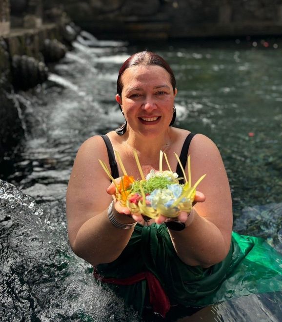 Woman in pink hat, seated in Balinese temple, hands in prayer, flanked by stone statues.