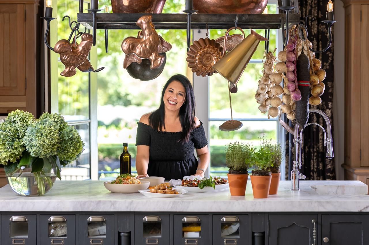 Woman smiles in a kitchen, standing behind a marble counter with food, herbs, and hanging copper cookware.