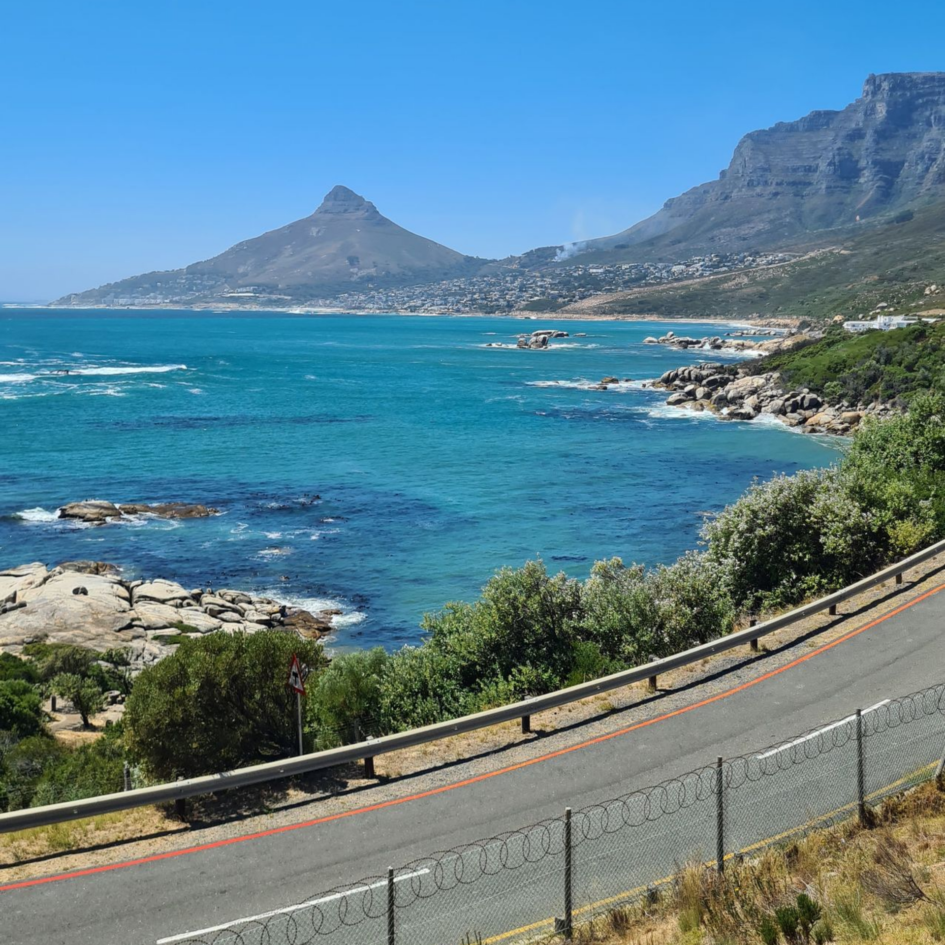 Coastal road with turquoise water, rocky shore, mountains, and blue sky.