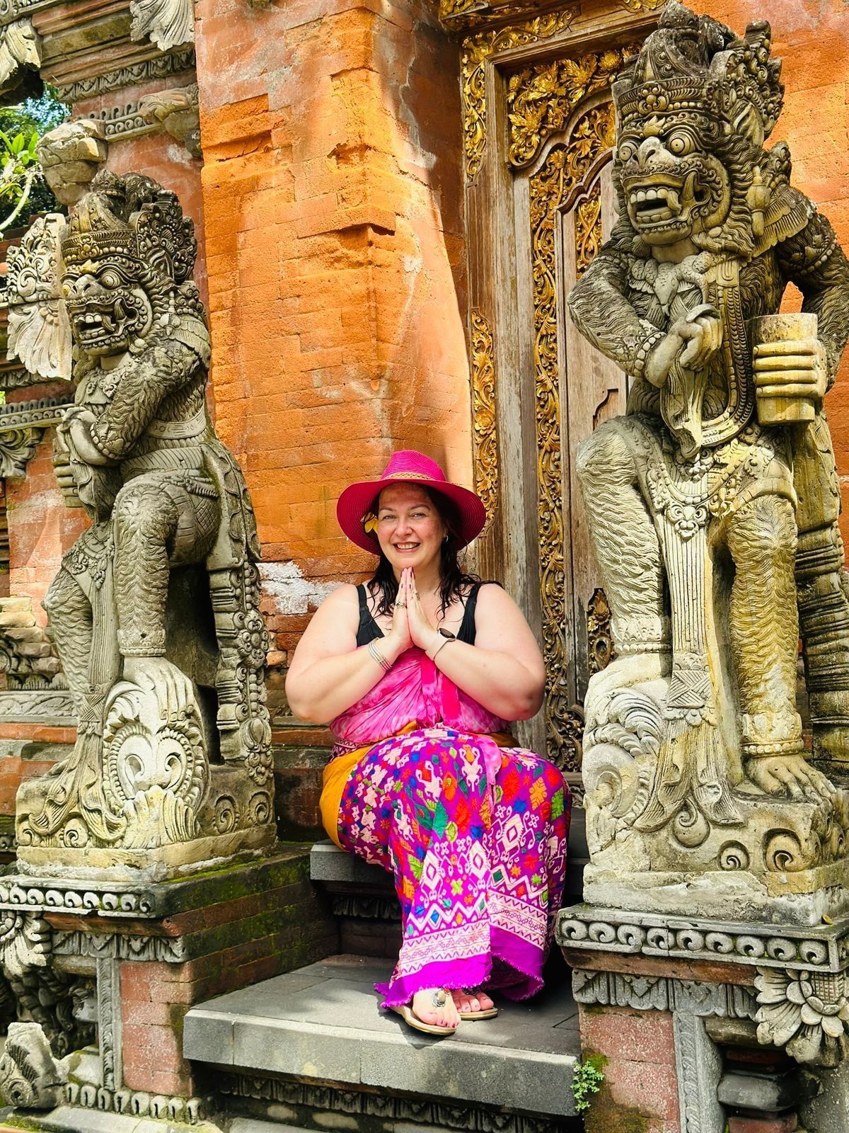 Woman in a pink hat and sarong, hands in prayer, sits between carved stone statues in a temple.