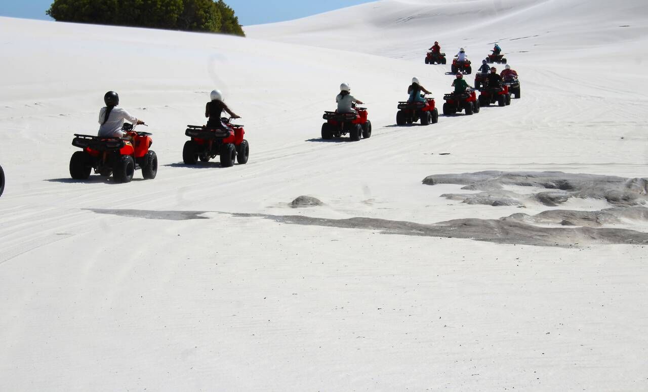 Quads' driving in a line across a white sand dune under a blue sky.