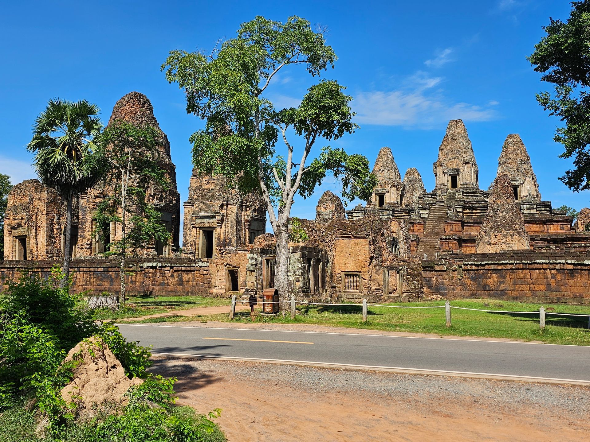 Ancient temple ruins with towers, tree in foreground, road, blue sky.