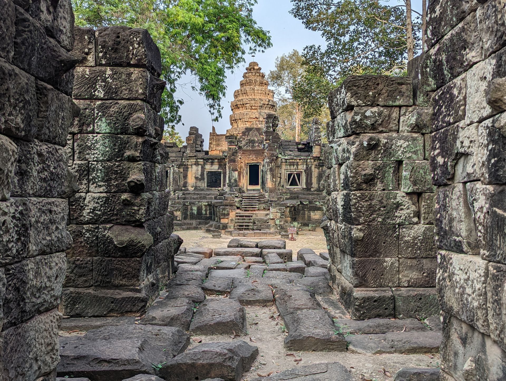 Stone ruins of a temple complex, viewed through a doorway, with trees and sky visible in the background.