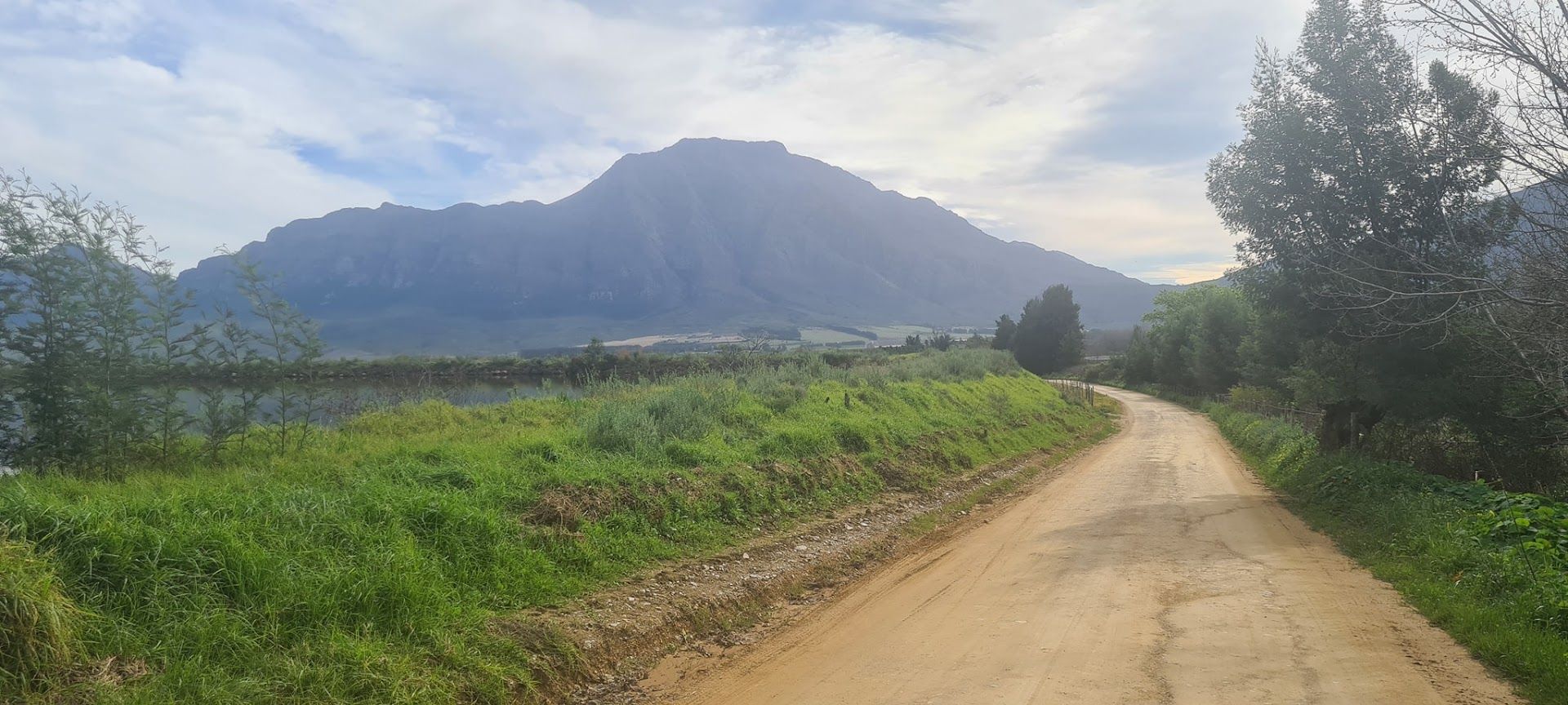 Dirt road leading towards a mountain, green grass on the side, and a cloudy sky.