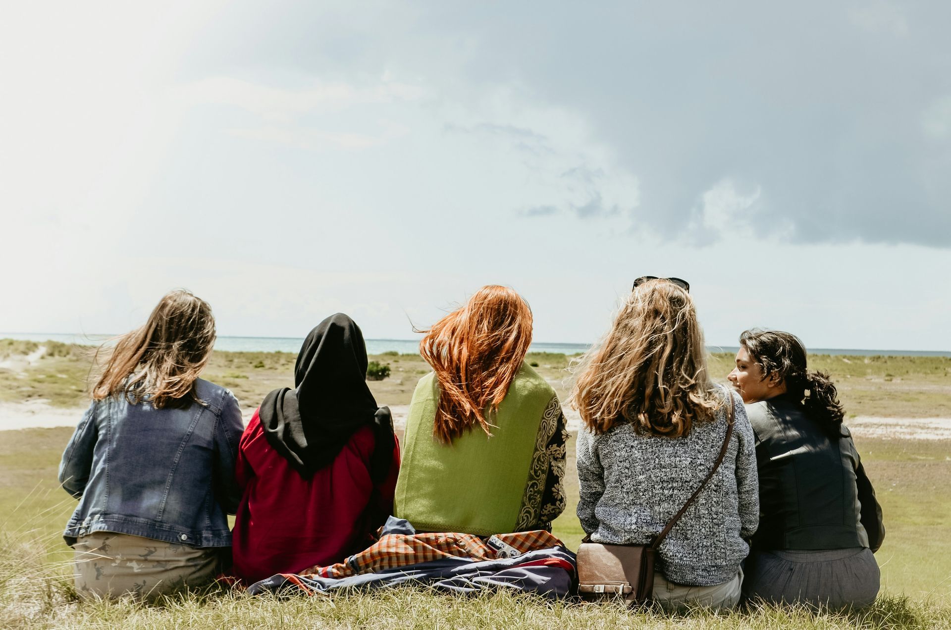Five people sit together on a grassy hill overlooking a beach, seen from behind, facing the ocean under a cloudy sky.