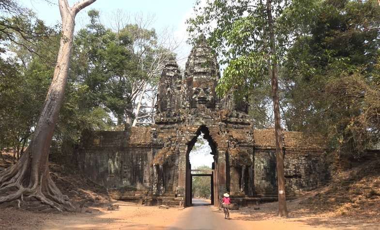 Stone gateway at Angkor Thom in Cambodia, weathered with trees and a person walking through.