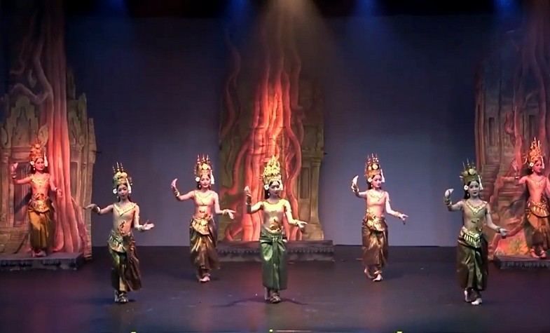 Apsara dancers on stage, wearing ornate headdresses, gold and green costumes. The background shows a temple and fire.
