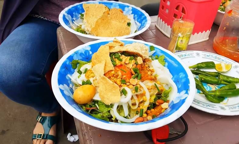 Bowls of Vietnamese noodles topped with shrimp, peanuts, and crackers, on a table with chili peppers and a woman wearing jeans.