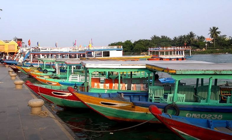 Boats docked at a pier; green, red, and yellow hulls; waterfront with buildings and trees.
