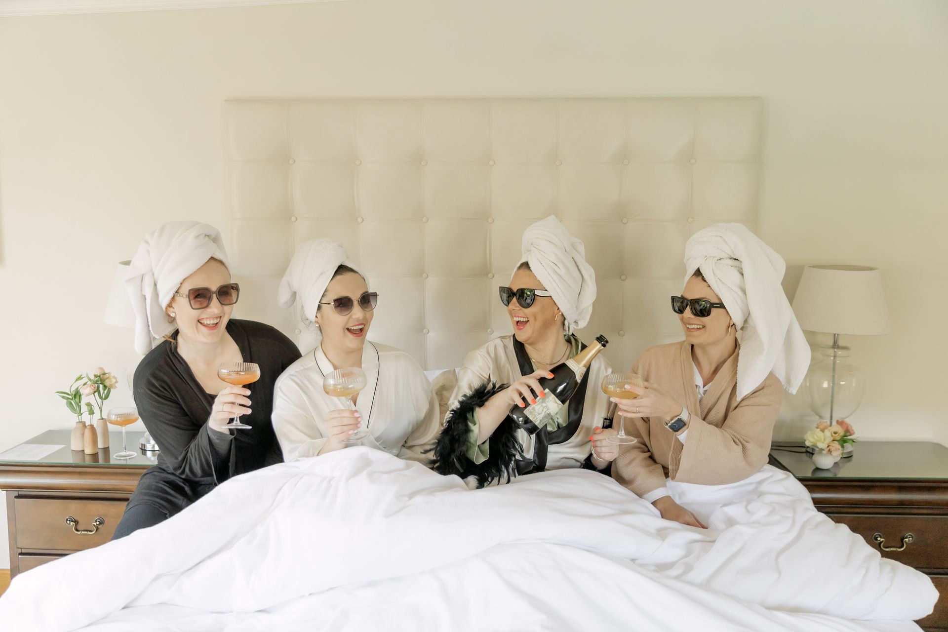 Four people wearing white hair towels and sunglasses toast with glasses of champagne while sitting on a bed.