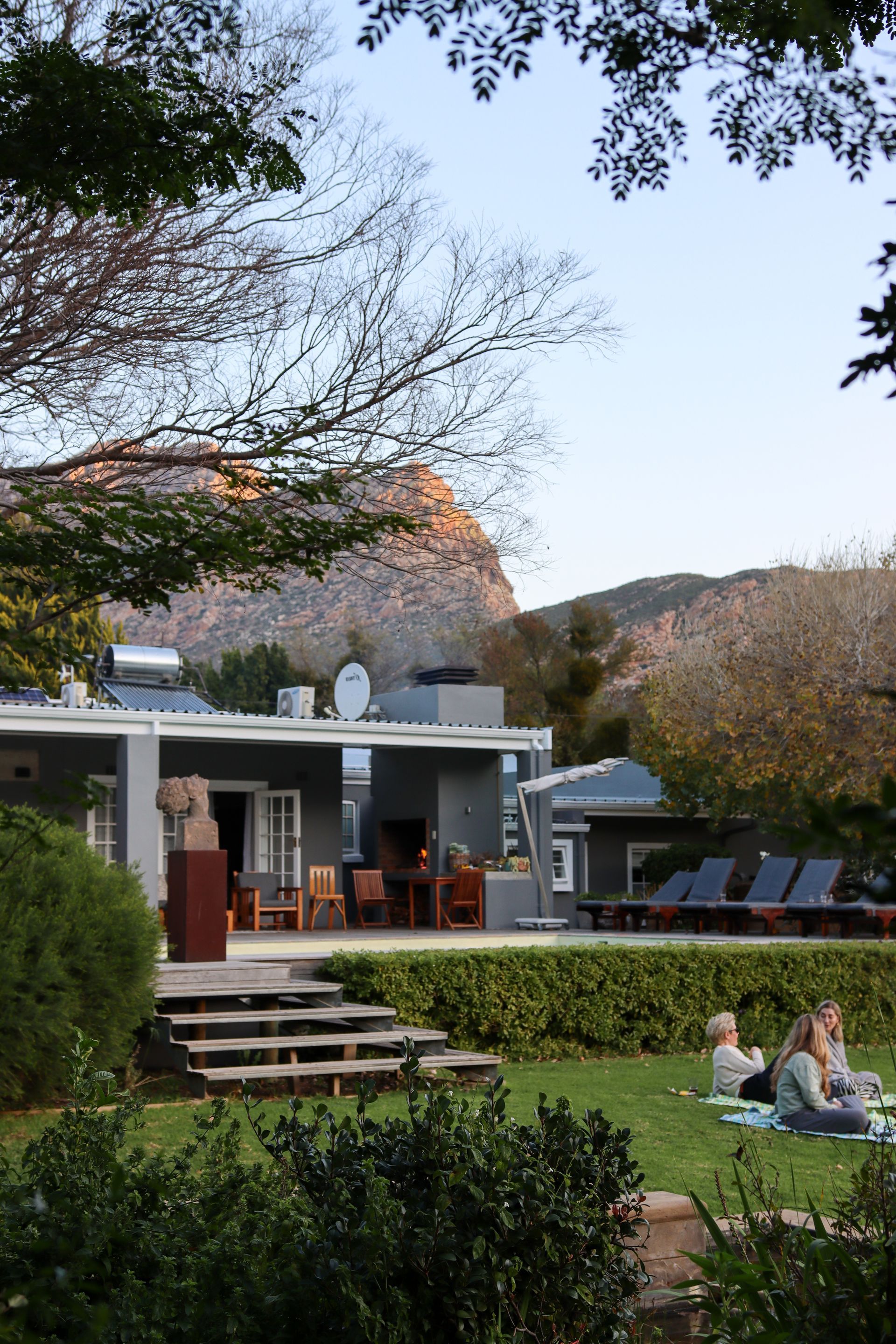 A grey house with a lawn and mountain backdrop. Two people sit on the grass.