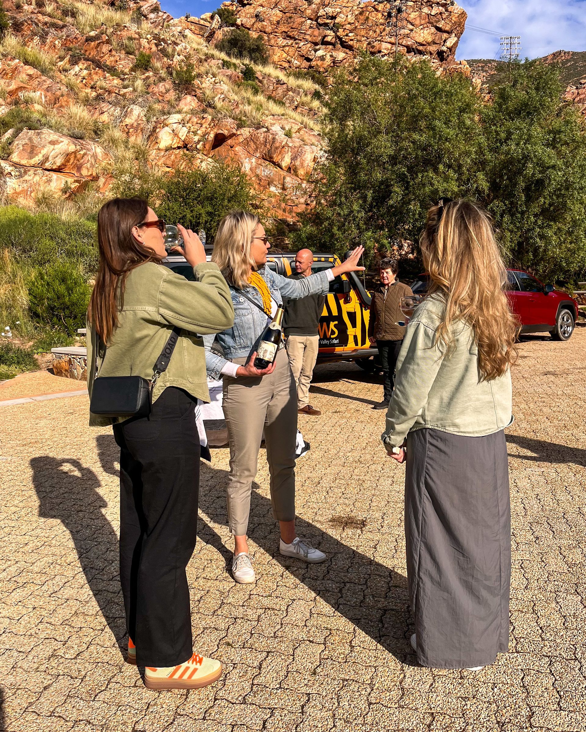 Three women talking outdoors near rocky hills; one points, others drink.