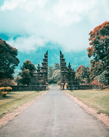 Stone gate entrance to a temple grounds. Paved road leads to the gates, surrounded by trees and misty mountains.