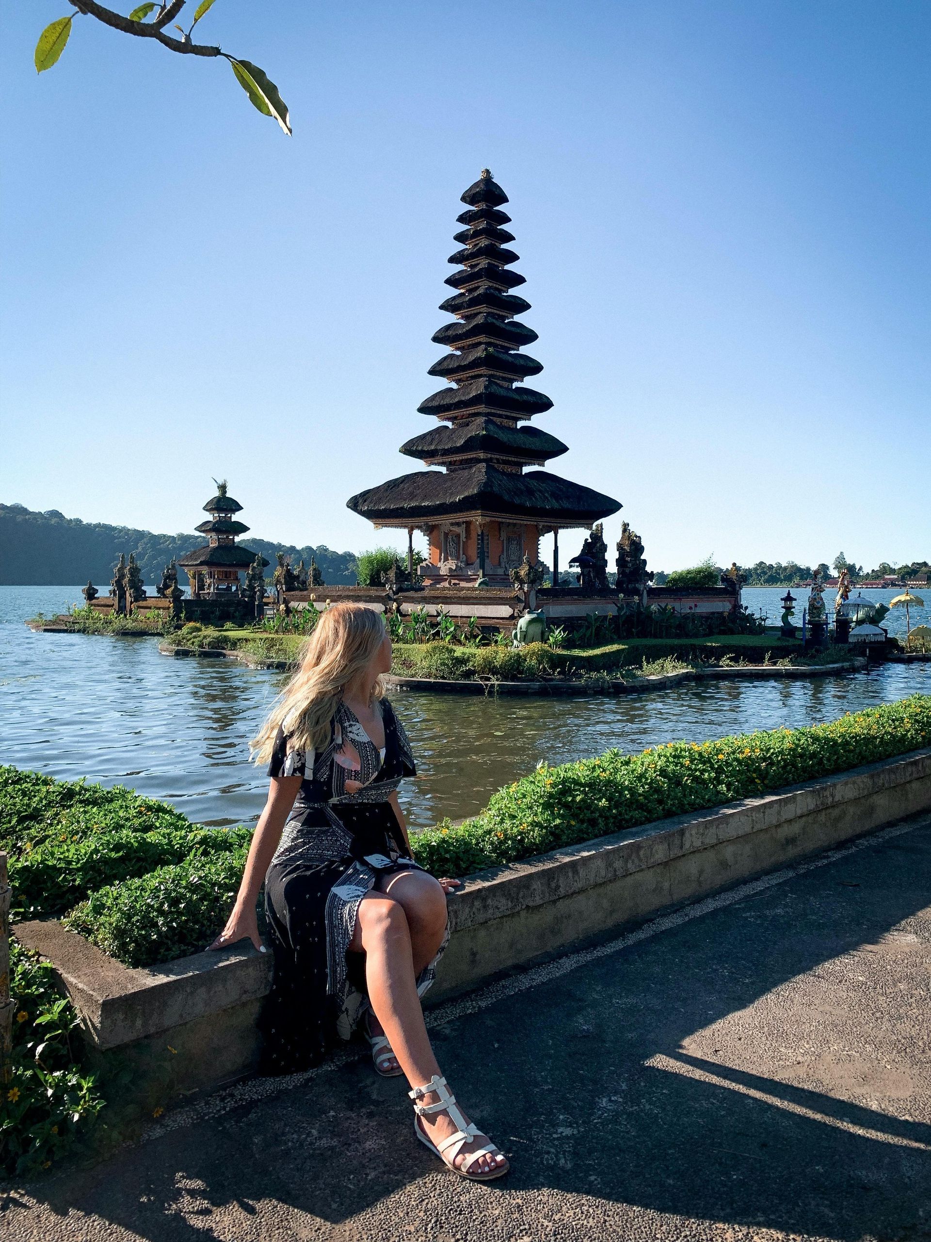 A woman sits on a stone wall overlooking Pura Ulun Danu Bratan, a tiered Hindu temple on a lake under a clear blue sky.