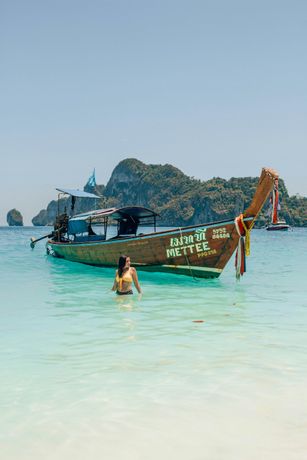 Woman wading in turquoise water near a longtail boat, mountains in background, sunny day.
