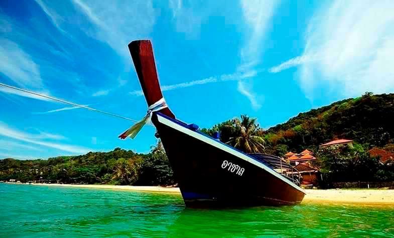 Longtail boat on a turquoise sea, sandy beach, lush green island in background, blue sky.