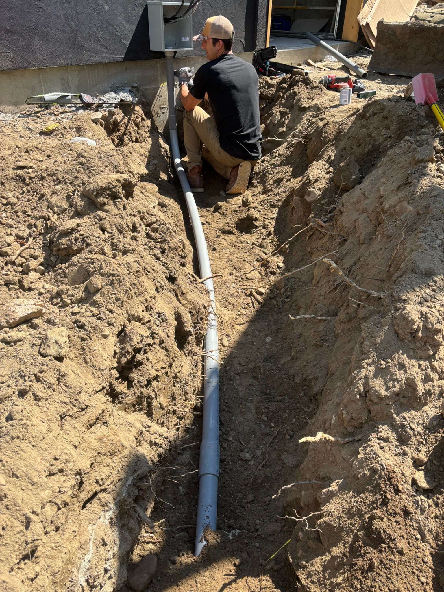 Man installs pipe in trench beside a building. Gray pipe, brown dirt, sunny outdoor setting.
